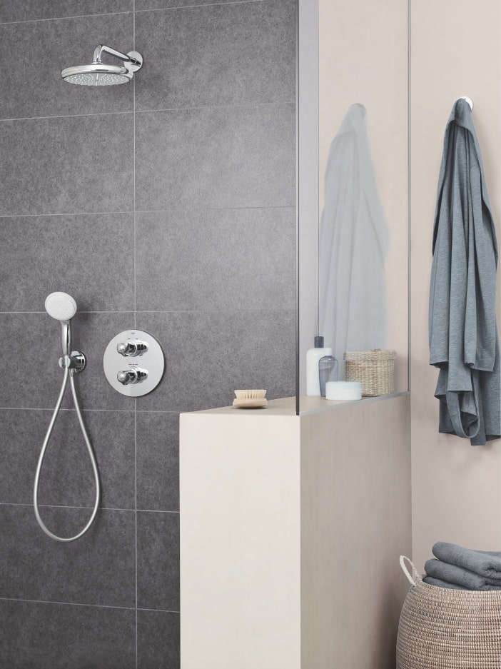 Prysznic z deszczownicą w łazience z ciemnymi kafelkami; obok półka z przyborami i zawieszony na jasnej ścianie ręcznik.